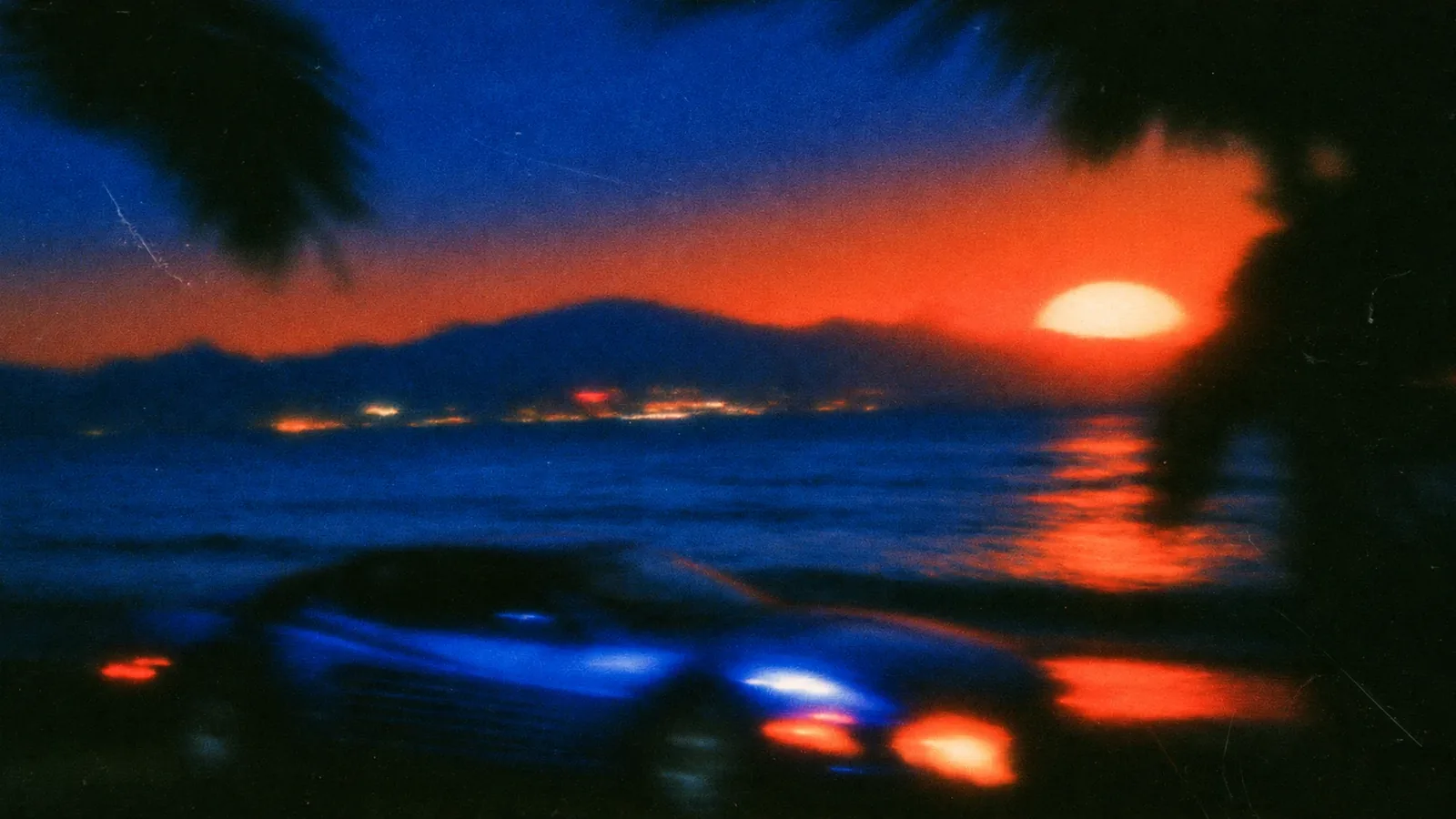 Grainy, soft-focus film still of a car parked by the sea at dusk, an orange sun setting behind dark mountains and palm fronds framing a cobalt-blue sky