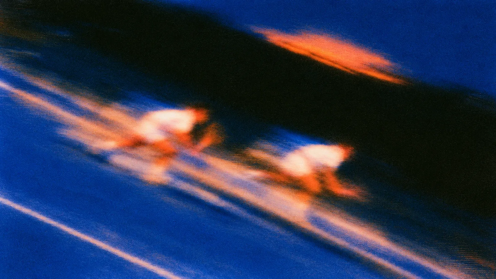 Grainy, motion-blurred blue and orange photo of two cyclists racing side by side along a lane, their shadow streaking dark across the track